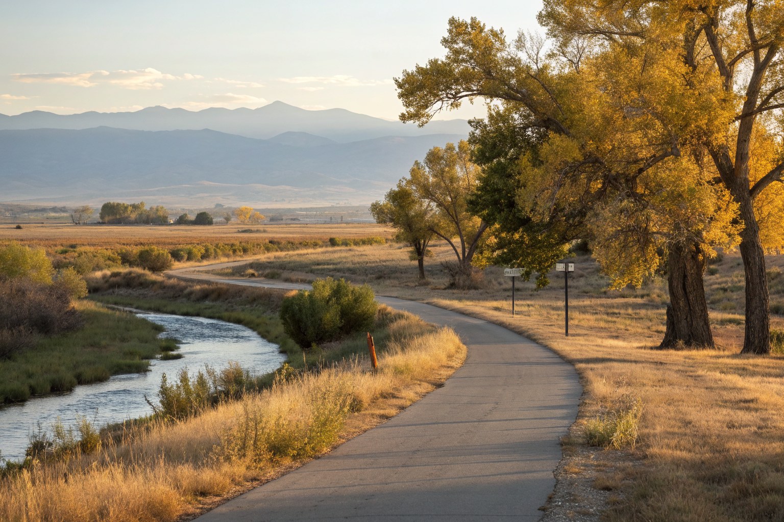 A paved trail curving beside a cottonwood-lined creek on the plains, mountains in the distance