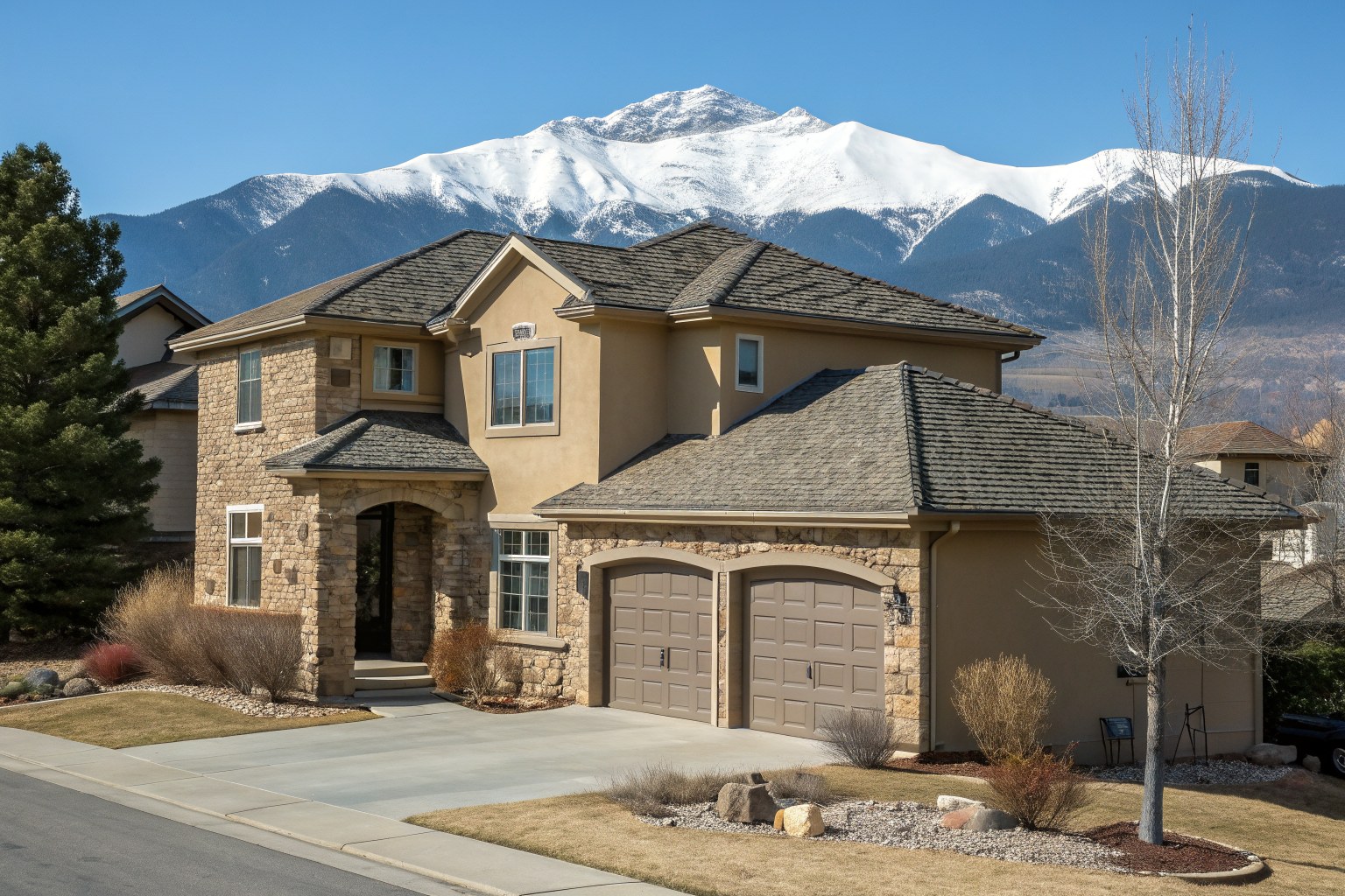 A two-story tan stucco and stone home with a charcoal shingle roof below a snow-capped mountain peak