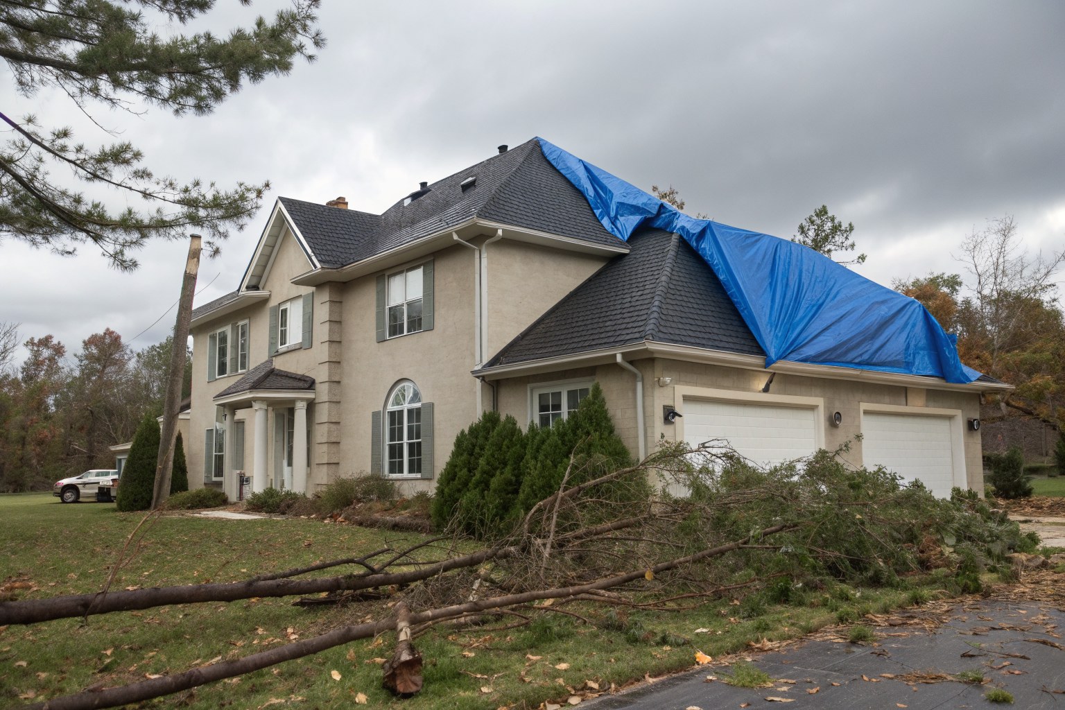 A two-story stucco house with a blue tarp over part of its roof and fallen pine limbs on the lawn